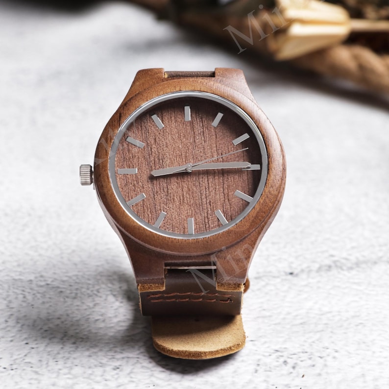 K&ouml;nnte beinhalten: Eine Holzarmbanduhr mit braunem Zifferblatt und silberner L&uuml;nette. Die Uhr hat ein braunes Lederarmband. Das Zifferblatt hat silberfarbene Stundenmarkierungen und Zeiger. Die Uhr ist aus Holz gefertigt und hat eine nat&uuml;rliche Maserung.