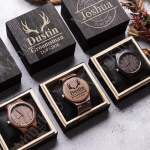 May include: Wooden watches and engraved boxes. The watch faces and boxes are engraved with names, dates, and a deer antler design. The watches are displayed on black cushions inside the boxes.