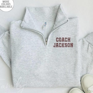 May include: A light gray quarter-zip sweatshirt with the text "COACH JACKSON" in maroon lettering. The sweatshirt has a collar and a zipper that extends down the chest. The words "MORE COLORS AVAILABLE" are in a white circle in the upper left corner.
