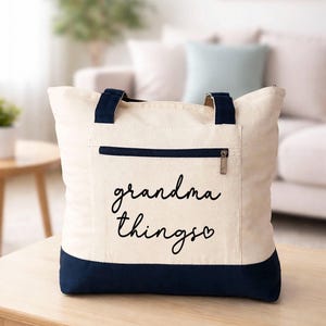 Pu&ograve; includere: Borsa tote color crema con dettagli blu navy. Ha una tasca frontale con cerniera e manici blu navy. Le parole "grandma things" sono scritte in corsivo nero sul davanti, con un simbolo a forma di cuore alla fine.