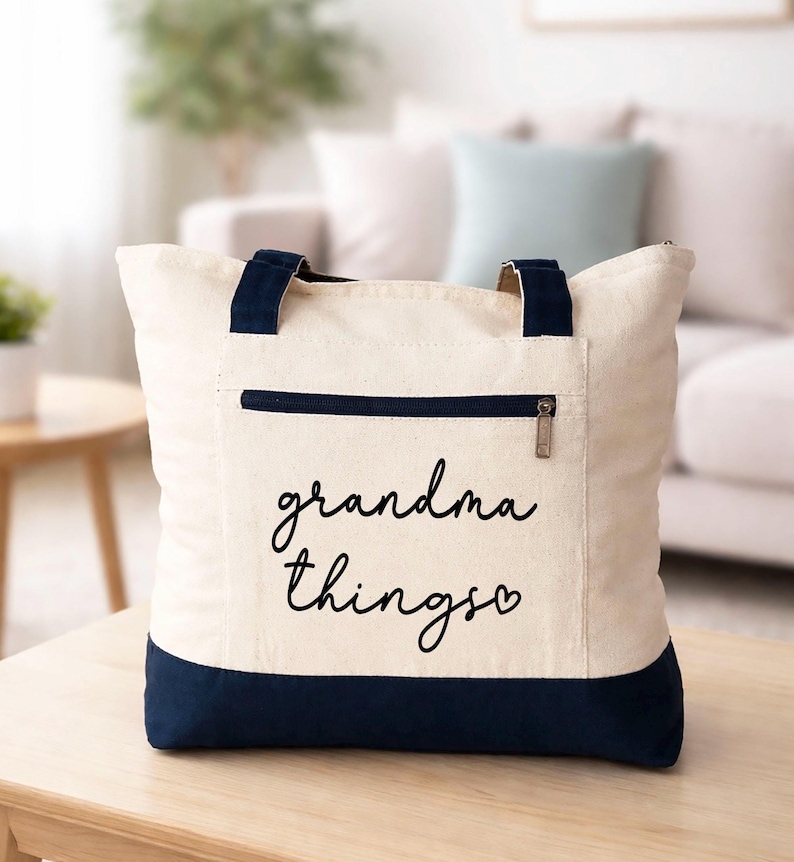 Pu&ograve; includere: Borsa tote color crema con dettagli blu navy. Ha una tasca frontale con cerniera e manici blu navy. Le parole "grandma things" sono scritte in corsivo nero sul davanti, con un simbolo a forma di cuore alla fine.