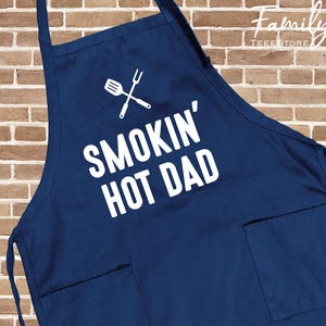 May include: A navy blue apron with white text reading "SMOKIN' HOT DAD" and a crossed spatula and fork graphic. The apron features two front pockets and adjustable neck and waist straps. The background is a brick wall.