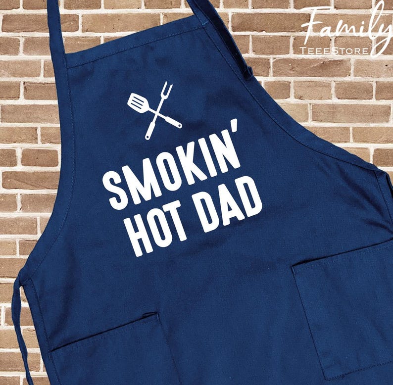 May include: A navy blue apron with white text reading "SMOKIN' HOT DAD" and a crossed spatula and fork graphic. The apron features two front pockets and adjustable neck and waist straps. The background is a brick wall.