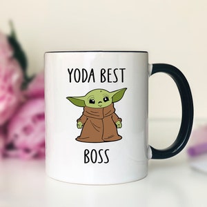Pu&ograve; includere: Tazza in ceramica bianca con manico e bordo neri. La tazza presenta un'illustrazione di un personaggio dei cartoni animati verde e il testo "YODA BEST BOSS" in maiuscolo nero. Lo sfondo &egrave; sfocato con fiori rosa.