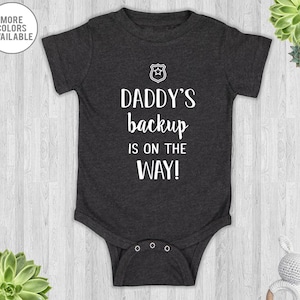 May include: A dark grey baby onesie with white text that reads "Daddy's backup is on the way!" and a small white police badge graphic.