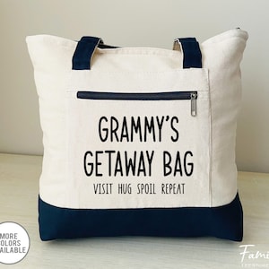 May include: A white canvas tote bag with black handles and a black bottom. The bag has a zippered pocket on the front with the text "Grammy's Getaway Bag Visit Hug Spoil Repeat" printed in black.