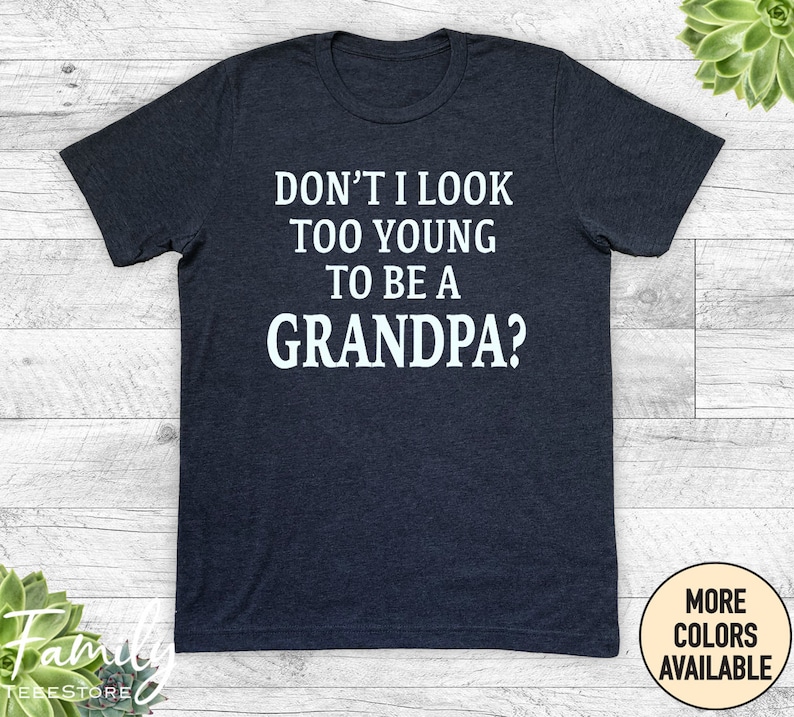 May include: A dark gray t-shirt with the text 'DON'T I LOOK TOO YOUNG TO BE A GRANDPA?' printed in white on the front. The t-shirt is displayed on a wooden surface. The text 'MORE COLORS AVAILABLE' is printed on a white tag in the bottom right corner of the image.