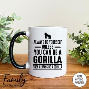 May include: White ceramic coffee mug with a black rim and handle. The mug features a black silhouette of a gorilla and the text "Always be yourself - unless - you can be a gorilla then always be a gorilla".