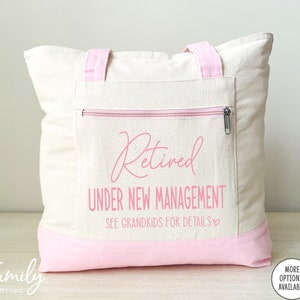 May include: A white canvas tote bag with pink handles and a pink zippered pocket. The bag has the text "Retired Under New Management See Grandkids for Details" printed on it in pink.