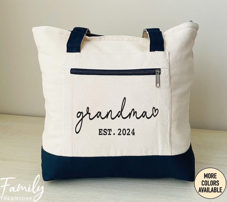 Puede incluir: Una bolsa de lona blanca con asas negras y fondo negro. La bolsa tiene un bolsillo con cremallera en la parte delantera y el texto "grandma EST. 2024" impreso en negro.