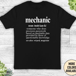 May include: Black t-shirt with white text that reads "mechanic" and a definition of the word. The definition includes the phonetic spelling, "muh-kan-ik", and a humorous description of the job. The text also includes "see also: wizard, magician".