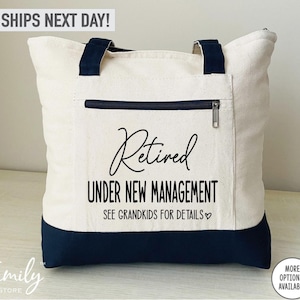 May include: A white canvas tote bag with black handles and a black zippered pocket. The bag has black text that reads "Retired Under New Management See Grandkids for Details".