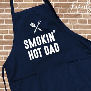 May include: A navy blue apron featuring the text "SMOKIN' HOT DAD" in white, along with a crossed spatula and fork graphic. It has two front pockets and neck and waist ties. The Family Tee Store logo is in the upper right corner.