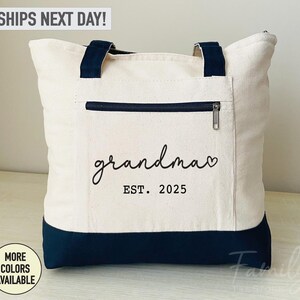 Puede incluir: Una bolsa de lona blanca con asas negras y fondo negro. La bolsa tiene un bolsillo con cremallera en la parte delantera y el texto "grandma est. 2025" impreso en negro.