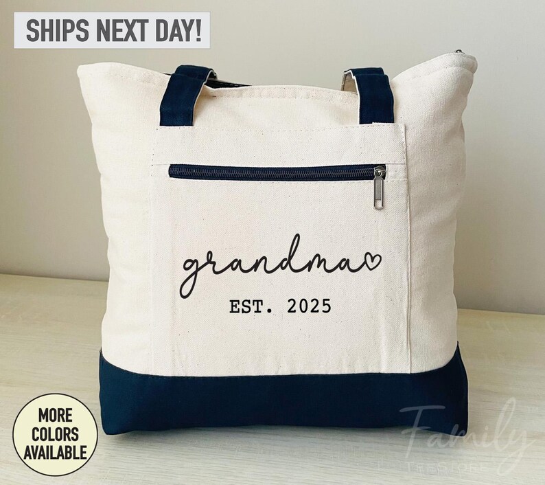 Puede incluir: Una bolsa de lona blanca con asas negras y fondo negro. La bolsa tiene un bolsillo con cremallera en la parte delantera y el texto "grandma est. 2025" impreso en negro.