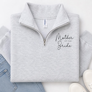 May include: A light gray quarter-zip sweatshirt featuring a silver zipper and ribbed cuffs. The text "Mother OF THE Bride" is printed in black script and block lettering on the upper right chest area. The garment includes a white inner neck label.