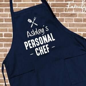 May include: A navy blue apron with the text "Ashley's PERSONAL CHEF" in white. The apron features a crossed spatula and whisk graphic above the text and two front pockets. The background is a brick wall.