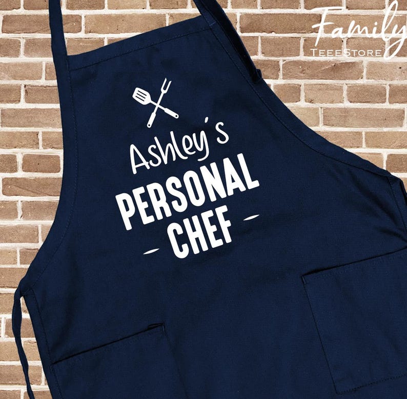 May include: A navy blue apron with the text "Ashley's PERSONAL CHEF" in white. The apron features a crossed spatula and whisk graphic above the text and two front pockets. The background is a brick wall.