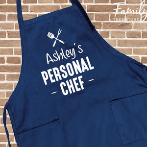 May include: A navy blue apron with white text that reads "Ashley's PERSONAL CHEF". The apron has two front pockets and a crossed spatula and fork graphic. The Family Tee Store logo is in the upper right corner.