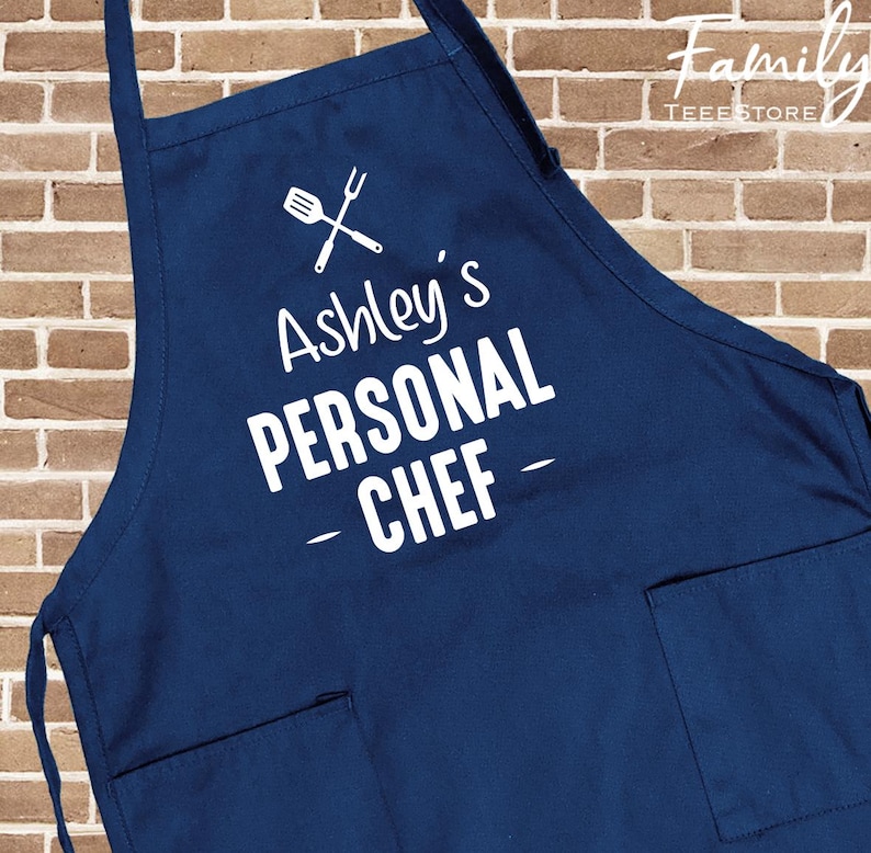 May include: A navy blue apron with white text that reads "Ashley's PERSONAL CHEF". The apron has two front pockets and a crossed spatula and fork graphic. The Family Tee Store logo is in the upper right corner.