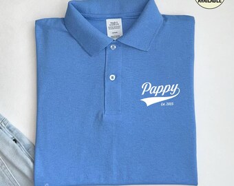 May include: A cornflower blue polo shirt featuring the word "Pappy" in white script with a white baseball-style banner, and "Est. 2025" underneath. The shirt has a button-down collar and is neatly folded.