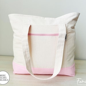 Puede incluir: Una bolsa de lona blanca con ribete rosa y un bolsillo con cremallera. La bolsa tiene dos asas largas.