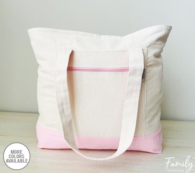 Puede incluir: Una bolsa de lona blanca con ribete rosa y un bolsillo con cremallera. La bolsa tiene dos asas largas.