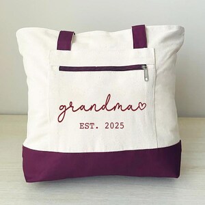 Puede incluir: Una bolsa de lona blanca con fondo morado. La bolsa tiene un bolsillo con cremallera y el texto "grandma EST. 2025" en rojo.