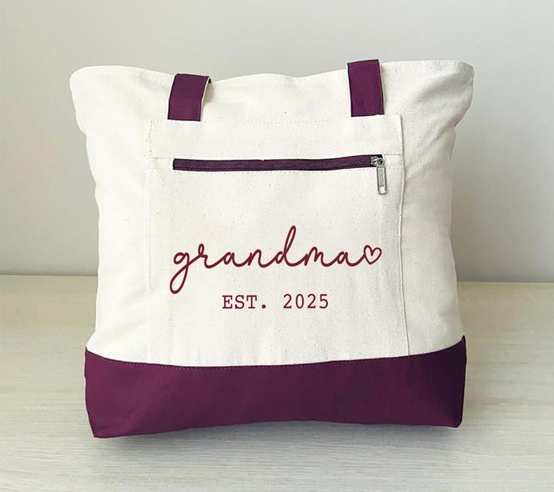 Puede incluir: Una bolsa de lona blanca con fondo morado. La bolsa tiene un bolsillo con cremallera y el texto "grandma EST. 2025" en rojo.