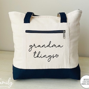 Pu&ograve; includere: Borsa tote in tela color crema con dettagli blu navy. La borsa ha una tasca frontale con cerniera e manici blu navy. Le parole "grandma things" sono stampate in corsivo nero sul davanti. Una borsa tote elegante e pratica.