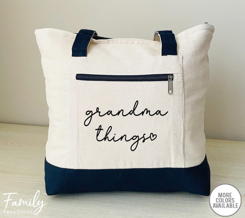 Pu&ograve; includere: Borsa tote in tela color crema con dettagli blu navy. La borsa ha una tasca frontale con cerniera e manici blu navy. Le parole "grandma things" sono stampate in corsivo nero sul davanti. Una borsa tote elegante e pratica.