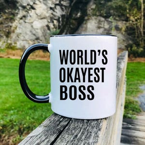 May include: White ceramic coffee mug with a black rim and handle. The mug has black text that reads "WORLD'S OKAYEST BOSS".