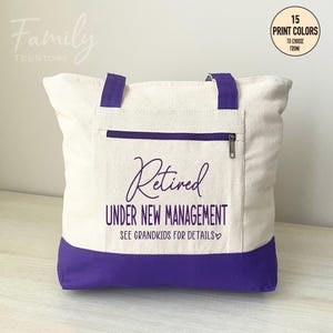 May include: A white canvas tote bag with purple handles and a purple bottom. The bag has a zippered pocket on the front and the text "Retired Under New Management See Grandkids for Details" printed in purple.