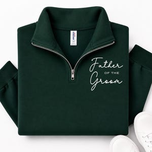 May include: A dark forest green quarter-zip sweatshirt featuring a silver metal zipper and a white script graphic on the chest that reads Father OF THE Groom. The garment has a high collar, long sleeves with ribbed cuffs, and a Jerzees brand label.