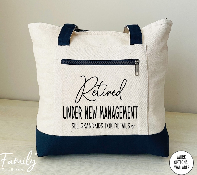 May include: A white canvas tote bag with black handles and a black bottom. The bag has a zippered pocket on the front and the text "Retired Under New Management See Grandkids for Details" in black.