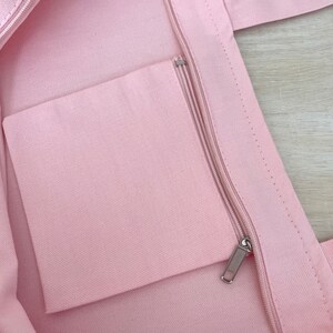 K&ouml;nnte beinhalten: Eine rosa Canvas-Tragetasche mit einer Rei&szlig;verschlusstasche im Inneren. Die Tasche hat einen Rei&szlig;verschluss und zwei Henkel.