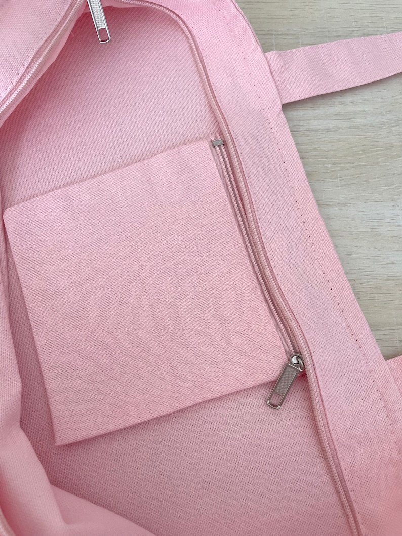 K&ouml;nnte beinhalten: Eine rosa Canvas-Tragetasche mit einer Rei&szlig;verschlusstasche im Inneren. Die Tasche hat einen Rei&szlig;verschluss und zwei Henkel.