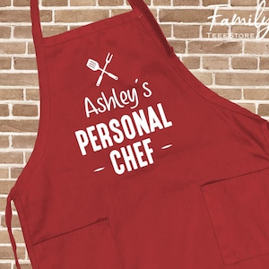 May include: A red apron with two front pockets and a neck strap. The apron features the text "Ashley's PERSONAL CHEF" in white, along with a graphic of a spatula and fork. The apron is made of a durable fabric.