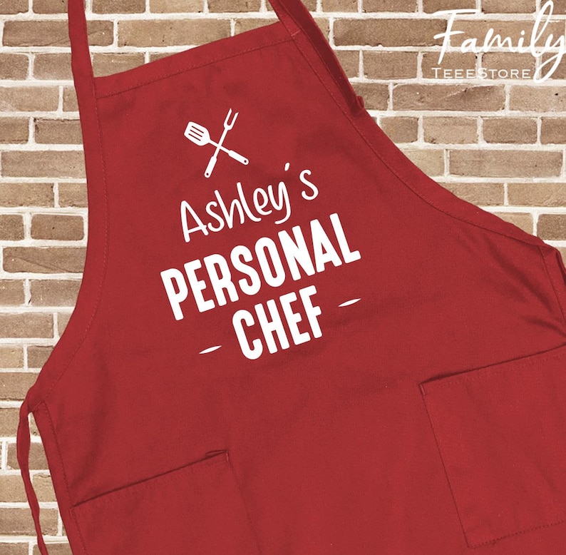 May include: A red apron with two front pockets and a neck strap. The apron features the text "Ashley's PERSONAL CHEF" in white, along with a graphic of a spatula and fork. The apron is made of a durable fabric.