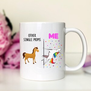 K&ouml;nnte beinhalten: Wei&szlig;e Keramik-Kaffeetasse mit einem humorvollen Design. Die linke Seite sagt "Other Single Moms" mit einem braunen Pferd. Die rechte Seite sagt "ME" mit einem Einhorn, das eine Stange h&auml;lt und einen Regenbogen-Schwanz hat. Der Hintergrund ist wei&szlig; mit bunten Sternen.