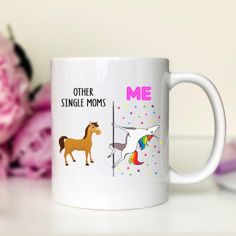 K&ouml;nnte beinhalten: Wei&szlig;e Keramik-Kaffeetasse mit einem humorvollen Design. Die linke Seite sagt "Other Single Moms" mit einem braunen Pferd. Die rechte Seite sagt "ME" mit einem Einhorn, das eine Stange h&auml;lt und einen Regenbogen-Schwanz hat. Der Hintergrund ist wei&szlig; mit bunten Sternen.