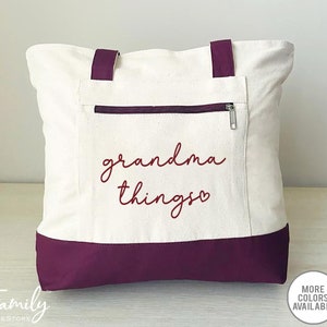 Pu&ograve; includere: Borsa tote color crema con base e manici viola scuro. La borsa presenta una tasca con cerniera e la scritta "grandma things" in corsivo bordeaux. Un piccolo cerchio indica che sono disponibili pi&ugrave; colori.