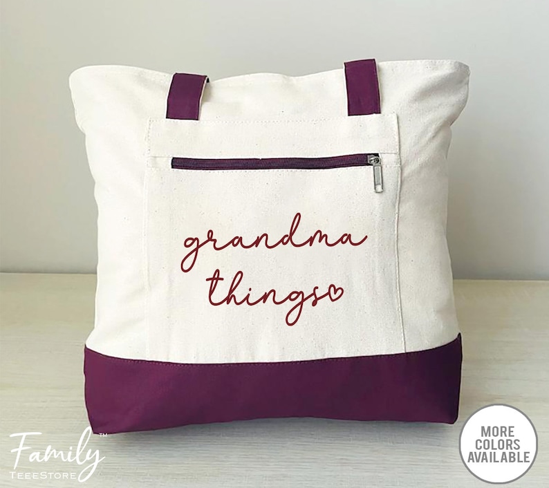Pu&ograve; includere: Borsa tote color crema con base e manici viola scuro. La borsa presenta una tasca con cerniera e la scritta "grandma things" in corsivo bordeaux. Un piccolo cerchio indica che sono disponibili pi&ugrave; colori.