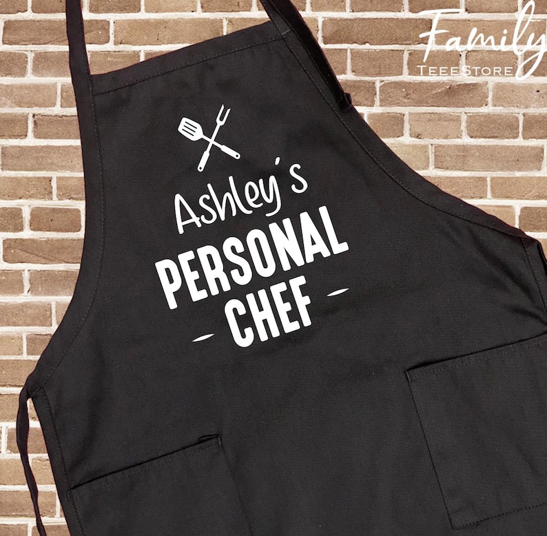 May include: Black apron with two front pockets and the text "Ashley's PERSONAL CHEF" in white. A graphic of a spatula and fork is above the text. The apron has a neck strap and waist ties.