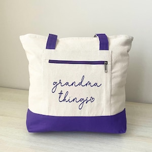 Pu&ograve; includere: Borsa tote color crema con dettagli viola. La borsa presenta un fondo e manici viola, una tasca con cerniera e la scritta "grandma things" in corsivo. Un accessorio pratico ed elegante.