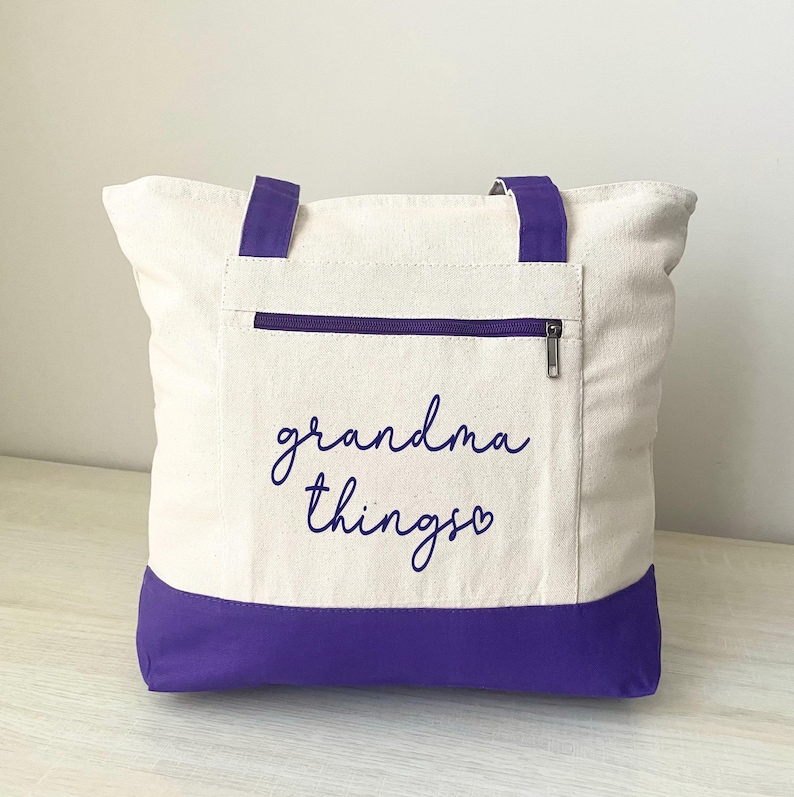 Pu&ograve; includere: Borsa tote color crema con dettagli viola. La borsa presenta un fondo e manici viola, una tasca con cerniera e la scritta "grandma things" in corsivo. Un accessorio pratico ed elegante.