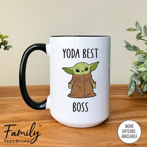 Pu&ograve; includere: Tazza in ceramica bianca con manico e bordo neri. La tazza presenta un'illustrazione a cartoni animati di un personaggio alieno verde con la scritta "YODA BEST BOSS". La tazza ha il testo "Family Teeestore" in basso.