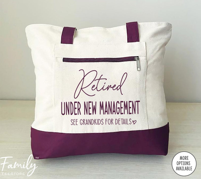 May include: A white canvas tote bag with maroon handles and a maroon bottom. The bag has a zippered pocket on the front and the text "Retired Under New Management See Grandkids for Details" printed in black.