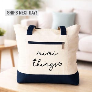 May include: Cream-colored tote bag with navy blue accents. The bag features a navy blue zipper pocket and handles. The words "mimi things" are written in black script on the front of the bag. The bag is sitting on a wooden table.