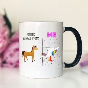 K&ouml;nnte beinhalten: Wei&szlig;e Keramik-Kaffeetasse mit schwarzem Henkel. Die Tasse hat eine schwarze Linie in der Mitte. Auf der linken Seite der Tasse steht der Text "Other Single Moms" mit einer Illustration eines braunen Pferdes. Auf der rechten Seite der Tasse steht der Text "ME" in Pink mit einer Illustration eines Einhorns, das eine Stange h&auml;lt.